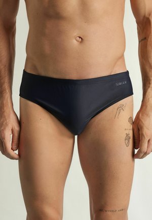 Męski tors w czarnych kąpielówkach z logo IUMAN, pokazujący tatuaże na prawej udzie i przedramieniu, na jednolitym jasnym tle.