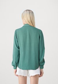 Camicia verde con texture, maniche lunghe, colletto e polsini con bottoni, abbinata a pantaloni corti bianchi sfilacciati. La vista da dietro mostra una vestibilità comoda.