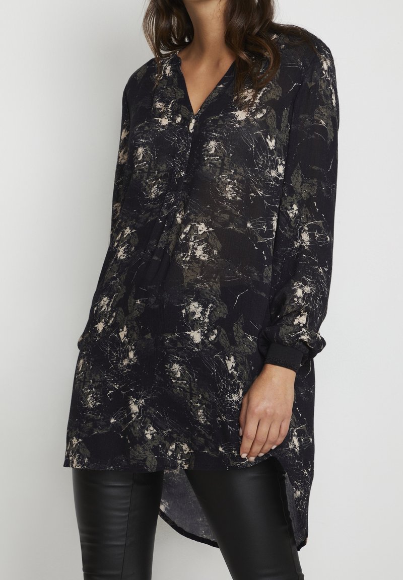 Femme portant une tunique noire à manches longues avec des motifs abstraits blancs et gris, associée à un pantalon en cuir noir, sur un fond uni.