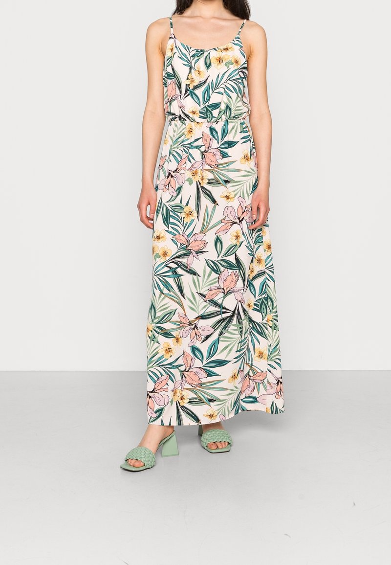 Femme portant une robe maxi sans manches à imprimé floral rose et jaune, associée à des sandales à talons blocs tissées vert clair.