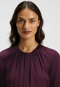 Burgunderfarbene Bluse mit plissiertem Ausschnitt, langen Ärmeln und glatter Stofftextur. Einfaches Design ohne sichtbare Muster oder Verzierungen.