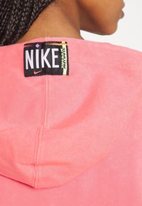 Růžová mikina s měkkou texturou. Na zadní části límce se nachází černá visačka Nike s nápisem "Just Do It" v žluté barvě.