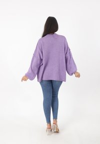 Lila übergroßer Pullover mit langen, weit geschnittenen Ärmeln, strukturiertem Strickstoff und Rundhalsausschnitt, kombiniert mit taillierten blauen Jeans und transparenten Absätzen.