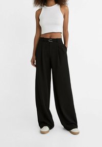 Pantalon noir à jambes larges avec plis, taille haute et détail de passants pour ceinture. Associé à un débardeur court blanc et des chaussures de couleur claire.