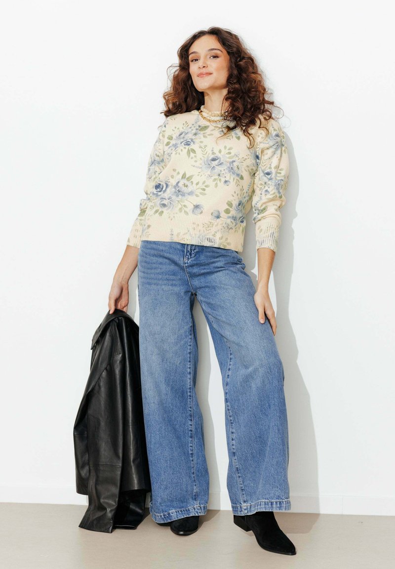 Femme portant un pull crème à fleurs, un jean bleu à jambes larges, des bottes noires, tenant une veste en cuir noire, debout contre un mur blanc.