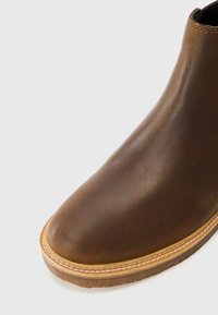 Brown leather ankle boot with a smooth surface, rounded toe, and contrasting beige stitching around the sole. Minimalist design.