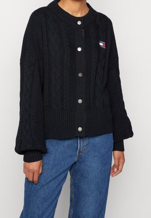 Personne portant un cardigan en maille torsadée noir avec des boutons et un patch logo Tommy Jeans, ainsi qu'un jean bleu, debout devant un fond uni.