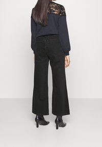 Mustat leveät farkut, joissa on korkea vyötärö, yhdistettynä tummansiniseen paitaan, jossa on pitsiä olkapäillä ja pitkät hihat. Korkeat mustat kengät.