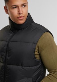 Schwarze gepolsterte Weste mit hohem Kragen, vorderem Reißverschluss und ohne Ärmel, getragen über einem olivgrünen Langarmshirt. Glatte Textur.