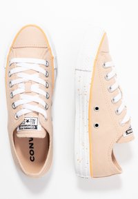 Baskets en toile beige avec des lacets blancs et des accents orange. Elles présentent une semelle en caoutchouc blanche texturée avec des détails tachetés et des œillets en métal.