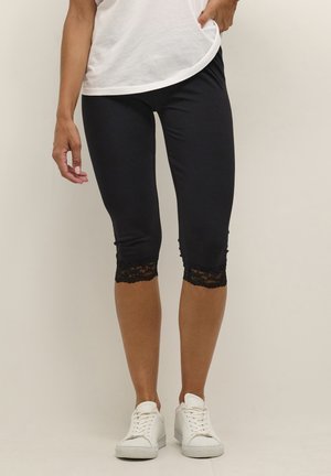 Black capri leggings made of stretchy fabric, featuring lace trim at the hem. Paired with white sneakers and a loose-fitting white shirt.
