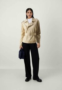 Geacă de piele bej cu talie cu curea, guler cu crestătură și închidere cu fermoar. Asortată cu pantaloni negri și o geantă shopper bleumarin.