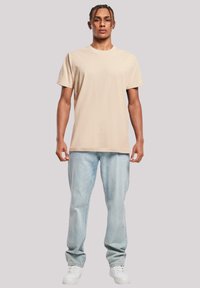 Beige cotton t-shirt with a crew neckline, paired with light blue denim jeans. Model wears white sneakers and has braided hair.
