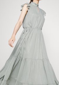 Ljusgrå maxi-klänning med veck och volangärmar, ett nyckelhålshalsringning och elastisk midja. Har en flerlagrad kjol och subtil glans.