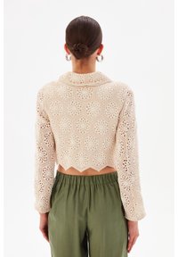 Maglione all'uncinetto beige con motivi esagonali e orlo scallop. Maniche lunghe con tagli intricati. Abbinato a pantaloni verdi con vita elastica.
