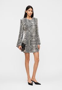 Femme portant une mini-robe manches longues à imprimé zèbre noir et blanc, tenant une pochette noire et chaussée de chaussures à talons noires.