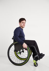 Zöld kiemelésekkel ellátott, fekete vázú kerekesszék, vékony fekete gumikerékkel. Ülő személy, aki sötétkék hosszú ujjú pólót és fekete nadrágot visel.