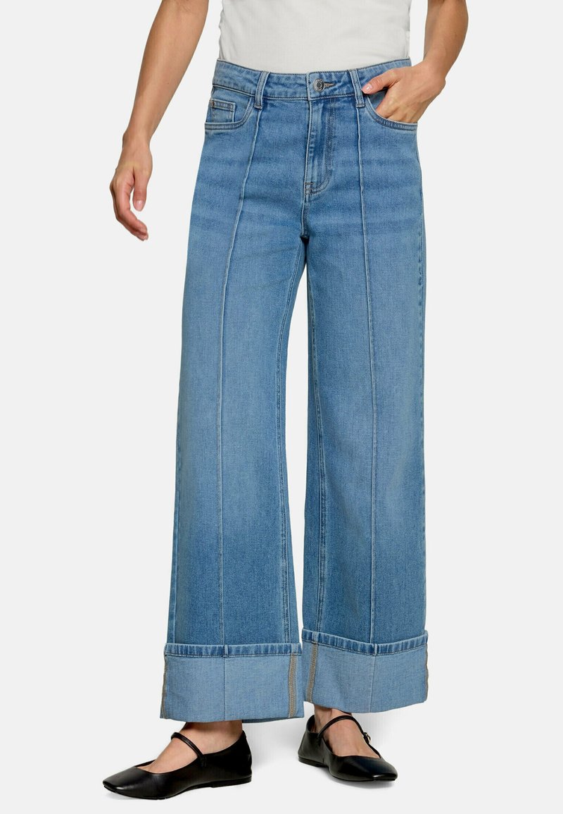 Personne portant un jean bleu clair à jambes larges avec des revers retroussés et des chaussures Mary Jane noires, debout sur un fond blanc.
