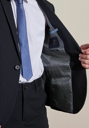 Uomo che indossa un abito scuro, camicia bianca e cravatta blu, che tiene aperta la giacca per mostrare la fodera grigia con motivo a piccoli puntini bianchi e l'etichetta interna.