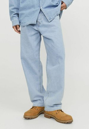 Personne portant un jean bleu clair ample, des bottes à lacets couleur beige et une chemise en denim bleu clair, avec une main dans la poche.