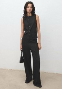 Top negru, fără mâneci, cu închidere cu buton și croială ajustată, purtat cu pantaloni largi. Modelul ține o geantă neagră în fața unui fundal simplu.