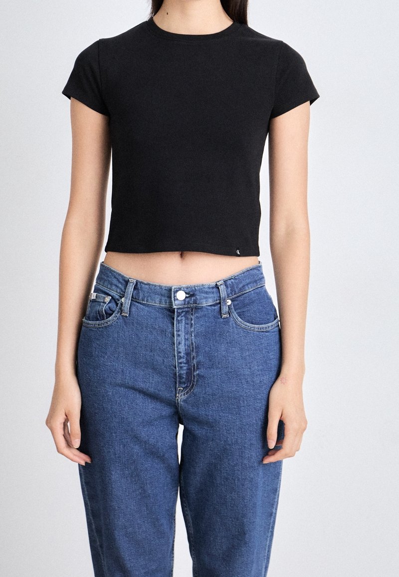 Jeune femme portant un haut court noir à manches courtes et un jean bleu taille haute, debout devant un fond uni.