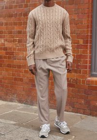 Pull beige en maille torsadée avec poignets côtelés, associé à un pantalon beige clair ajusté et des baskets grises à lacets blancs. Fond en briques texturées.