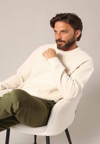 Crème trui met een geribde halslijn en textuurstof, gecombineerd met olijfgroene broek, zittend in een witte, pluchen stoel.