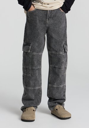 Person iført sorte vaskede cargobukser med for- og sidelommer, beige slip-on sko, hænder i lommerne, stående på en grå baggrund.