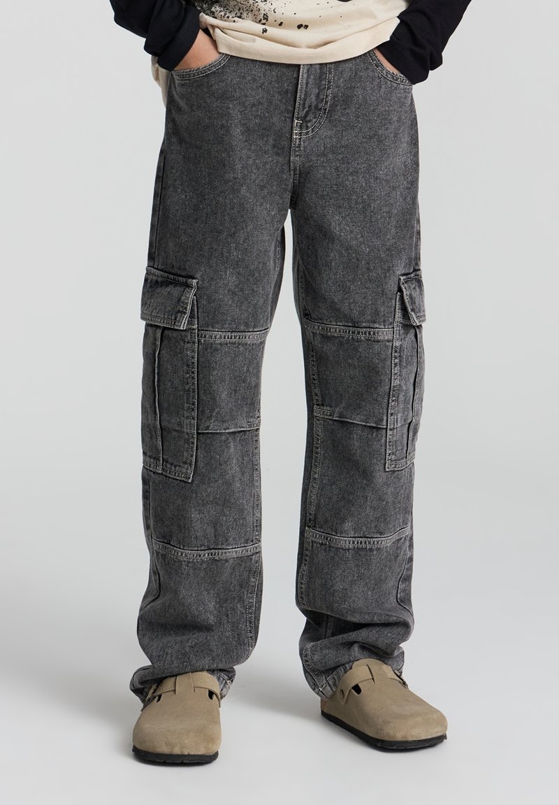 Personne portant un pantalon cargo noir délavé avec poches avant et latérales, chaussures beiges sans lacets, mains dans les poches, debout sur un fond gris.
