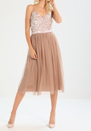 Femme portant un top sans manches en sequins or rose avec une jupe midi en tulle marron clair et des talons pointus couleur nude, posant avec une main près du visage.