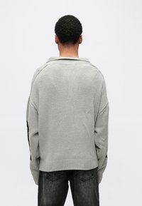 Ανοιχτό γκρι πουλόβερ με ριμπ τελείωμα και γιακά σαν μπολερό· διαθέτει κατεβασμένους ώμους και υφή στο ύφασμα. Μαύρες λεπτομέρειες στα μανίκια.