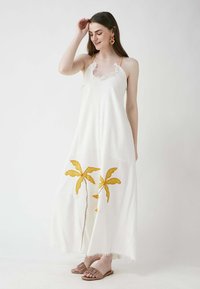 Weißes Maxikleid mit gelber Palmbaumstickerei, Spaghetti-Trägern und Fransenverzierungen am Saum, kombiniert mit beigen Rutsch-Sandalen.