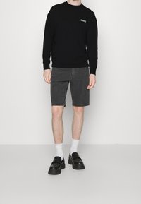 Felpa nera con un piccolo logo, abbinata a pantaloni short grigio scuro. Le calzature includono scarpe slip-on nere e calze bianche alla caviglia.