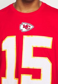Czerwona koszulka z białym, perforowanym numerem "15" obramowanym na żółto, z wyszywaną logo Kansas City Chiefs powyżej.