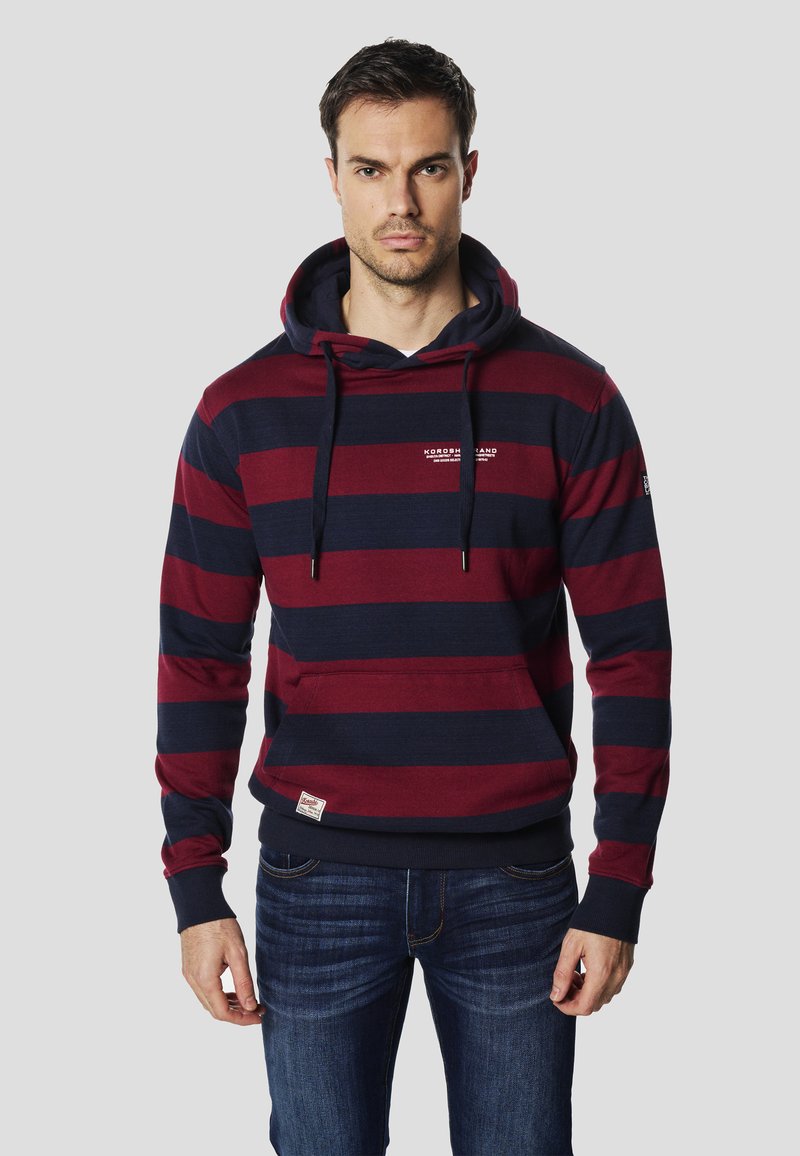 Bordovo-modrá pruhovaná mikina s kapucňou, predným vreckom, nastaviteľným šnúrkou a malým logom na hrudi, v kombinácii s tmavými džínami.