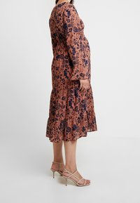 Gravid kvinna klädd i en långärmad, blommönstrad midikjol och beige band sandaletter med klack, som står mot en enfärgad bakgrund.