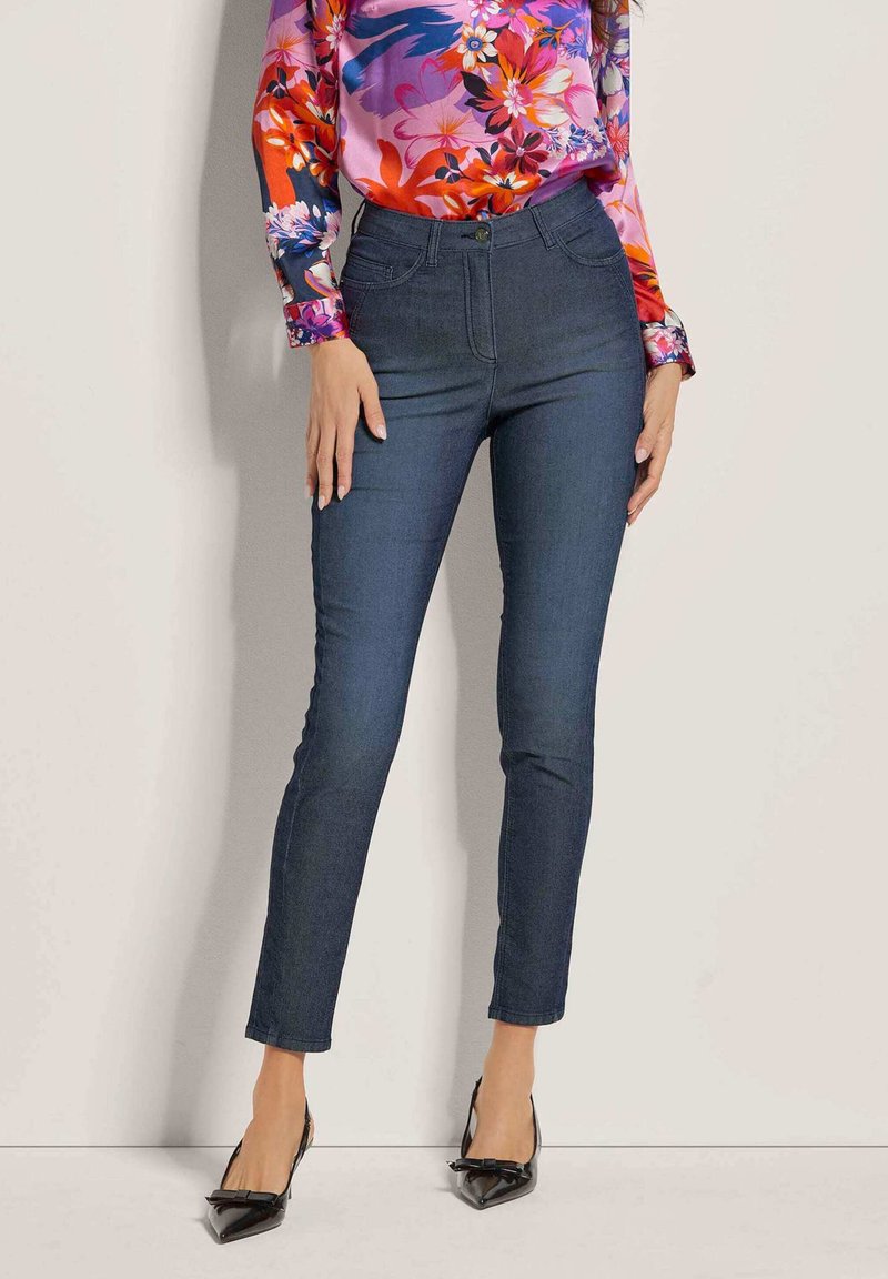 Donkerblauwe denim jeans met een slim fit, voorzien van voorzakken, een knoopsluiting, en gecombineerd met een blouse met bloemenpatroon en zwarte hakken.