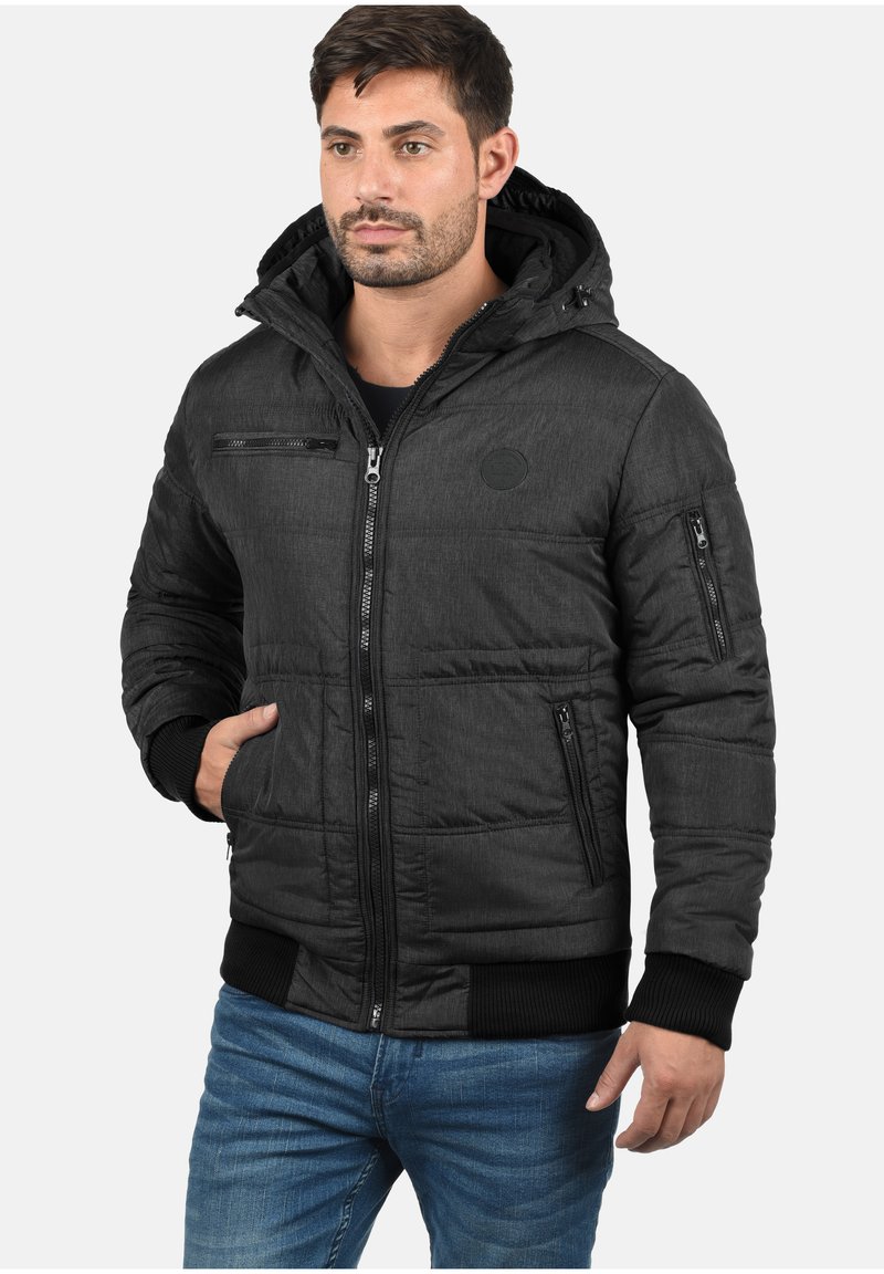 Schwarze, wattierte Jacke mit Kapuze, ausgestattet mit einer Reißverschlusstasche auf der Brust und Seitentaschen. Gerippte Bündchen und Saum, strukturierter Stoff.