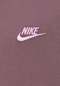 Μωβ υφασμάτινη μπλούζα με κεντημένο ροζ λογότυπο Nike στην αριστερή πλευρά. Το υλικό φαίνεται μαλακό και με υφή, με διακριτικό σχέδιο.