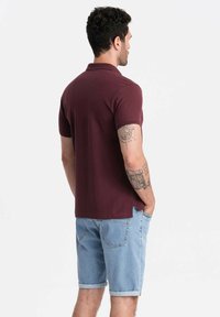 Burgundin lyhythihainen polo-paita, jossa on kaulus, yhdistetty vaaleansinisiin farkkushortseihin, joissa on käänteet. Kankaan näyttää olevan sileä; siinä on sivuihin leikkaukset.