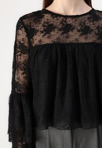 Blusa nera con una sezione inferiore strutturata e plissettata e un tessuto superiore trasparente ricamato a fiori. Maniche a campana lunghe.