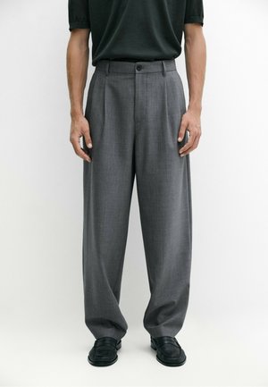 Mand iført løse grå bukser, sort bælte, sorte loafers og en mørk kortærmet skjorte står foran en ensfarvet hvid baggrund.