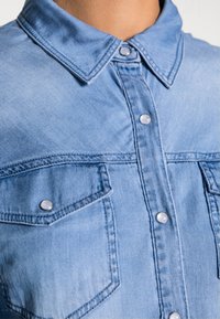 Camicia in denim azzurro chiaro con colletto a punta, bottoni a pressione argentati e due tasche sul petto con pattine. Il tessuto ha una finitura morbida e testurizzata.