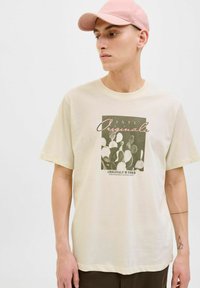 Krémové bavlněné tričko s grafikou kaktusu a textem "INTL Originals © 1990." Model má na sobě světle růžovou čepici. Krátké rukávy, volný střih.