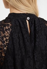 Schwarzes Spitzenoberteil mit hohem Kragen, Schlüsselanierschnitt hinten und zwei Knöpfen. Der Stoff hat florale Muster und eine strukturierte Oberfläche.