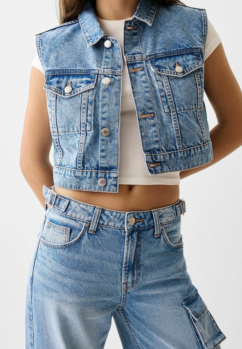 Světle modrá denimová vesta bez rukávů, s náprsními kapsami a viditelným šitím. V kombinaci se světle modrými džíny s vysokým pasem.