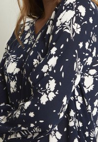 Gros plan sur une personne portant un chemisier bleu marine à motifs floraux blancs et un col en V, montrant le haut du torse et une partie du bras.