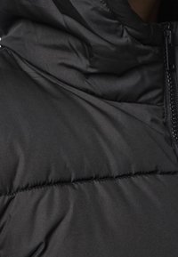 Svart puffjacka i närbild, med quiltade sektioner, en slät matt yta, hög krage och en sidodragningsdetalj.