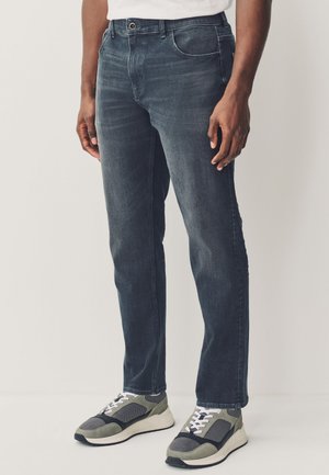 Mężczyzna w ciemnoszarych dopasowanych dżinsach i szaro-białych trampkach stoi na jasnym tle.